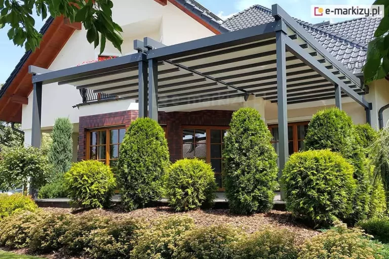 Dwie pergole Solid Aluprof ustawione na dużym tarasie przy piętrowym domku ze spadzistym dachem. Solidne aluminiowe konstrukcje w kolorze antracytu oraz biała tkanina dachowa komponują się z otoczeniem tarasu, zarówno na fasadzie budynku, jak i ogrodem pełnym zielonych krzewów wiecznozielonych oraz innych kwitnących roślin.