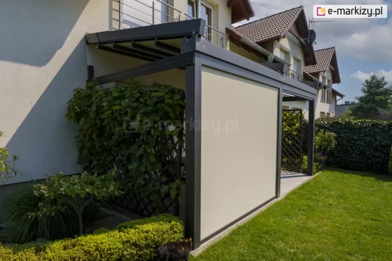 Zdjęcie prezentuje nowoczesną pergolę tarasową model Solid Aluprof w kolorze antracytowym, zamontowaną do ściany jasnego budynku jednorodzinnego. Głównym elementem wyróżniającym tę realizację jest pionowa przesłona boczna – refleksol przeciwsłoneczny Ziiip Selt w kolorze kremowym, który jest całkowicie opuszczony, wypełniając przestrzeń między słupami konstrukcyjnymi. Zadaszenie pergoli wykonane z segmentowej tkaniny jest częściowo zwinięte w stronę elewacji. Po lewej stronie konstrukcji widoczna jest drewniana kratka porośnięta gęstymi, zielonymi pnączami, a u podstawy znajduje się niski, zadbany żywopłot. Przed pergolą rozciąga się zielony trawnik. W tle widoczne są inne budynki mieszkalne z brązowymi dachami oraz błękitne niebo z lekkimi chmurami. Całość instalacji tworzy osłoniętą i prywatną przestrzeń wypoczynkową na tarasie.