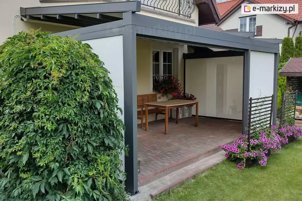 Nowoczesna pergola tarasowa o antracytowej, aluminiowej konstrukcji przyściennej, osadzona na podwyższonym tarasie wykonanym z czerwono-brązowej kostki brukowej. Konstrukcja posiada system bocznych przesłon typu screen w kolorze białym, które są częściowo opuszczone na bocznych ścianach, tworząc półotwartą przestrzeń. Zadaszenie pergoli składa się z ciemnych profili, nad którymi widoczny jest rozwinięty jasny materiał osłonowy. Wewnątrz pergoli, pod oknem budynku, ustawiono drewniany stół z jasnym blatem oraz dopasowaną drewnianą ławkę. Parapet okienny zdobią donice z obficie kwitnącymi czerwonymi kwiatami. Bezpośrednio przed wejściem do pergoli, na trawniku, znajdują się dwie niskie, czarne kratki ogrodowe, przy których rosną intensywnie różowe kwiaty. Po lewej stronie kadru dominuje wysoki, gęsty krzew o zielonych liściach, niemal całkowicie zasłaniający jeden ze słupów konstrukcyjnych. W tle widoczny jest fragment sąsiedniego domu z białymi ścianami i czerwonym dachem oraz zielone tuje wyznaczające granicę posesji.