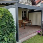 Nowoczesna pergola tarasowa o antracytowej, aluminiowej konstrukcji przyściennej, osadzona na podwyższonym tarasie wykonanym z czerwono-brązowej kostki brukowej. Konstrukcja posiada system bocznych przesłon typu screen w kolorze białym, które są częściowo opuszczone na bocznych ścianach, tworząc półotwartą przestrzeń. Zadaszenie pergoli składa się z ciemnych profili, nad którymi widoczny jest rozwinięty jasny materiał osłonowy. Wewnątrz pergoli, pod oknem budynku, ustawiono drewniany stół z jasnym blatem oraz dopasowaną drewnianą ławkę. Parapet okienny zdobią donice z obficie kwitnącymi czerwonymi kwiatami. Bezpośrednio przed wejściem do pergoli, na trawniku, znajdują się dwie niskie, czarne kratki ogrodowe, przy których rosną intensywnie różowe kwiaty. Po lewej stronie kadru dominuje wysoki, gęsty krzew o zielonych liściach, niemal całkowicie zasłaniający jeden ze słupów konstrukcyjnych. W tle widoczny jest fragment sąsiedniego domu z białymi ścianami i czerwonym dachem oraz zielone tuje wyznaczające granicę posesji.