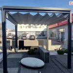 Wolnostojąca pergola tarasowa Tako o konstrukcji aluminiowej w kolorze antracytowym, z zadaszeniem z szarej tkaniny rzymskiej, ustawiona na zewnętrznym placu ekspozycyjnym. Materiał dachu jest rozwinięty, tworząc regularne, trapezowe zakładki (fałdy) podwieszone na poprzecznych profilach. Pergola oparta jest na czterech pionowych słupach o kwadratowym przekroju, zamontowanych na podłożu z ciemnych płyt tarasowych i kostki brukowej. Wewnątrz pergoli znajduje się biały, owalny element dekoracyjny przypominający kamień oraz niska roślinność w żwirze. W tle widoczny jest parking z samochodami, duża ciężarówka z naczepą oraz fragmenty budynków handlowych z czerwonymi akcentami i logotypami. Niebo jest bezchmurne, błękitne, a oświetlenie wskazuje na słoneczny dzień.