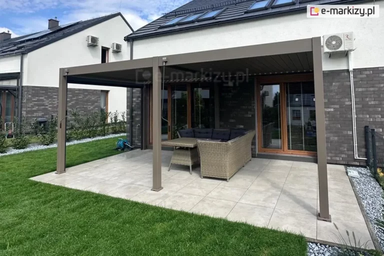 Szeroka, dwumodułowa pergola tarasowa w kolorze taupe, zamontowana jako konstrukcja przyścienna do elewacji nowoczesnego domu. Zadaszenie opiera się na trzech aluminiowych słupach frontowych i składa się z dwóch niezależnie sterowanych sekcji lamelowych, które w tym ujęciu pozostają w pozycji całkowicie zamkniętej. Pod pergolą, na tarasie wyłożonym jasnoszarymi płytami gresowymi, znajduje się narożny zestaw mebli ogrodowych z technorattanu z ciemnymi poduszkami oraz prostokątny stół. Przy dwóch słupach widoczne są mechanizmy korbowe do manualnej obsługi dachu. Elewacja budynku łączy ciemną cegłę klinkierową na poziomie parteru z jasnym tynkiem na piętrze, a na dachu widoczne są liczne okna połaciowe. Po lewej stronie znajduje się zadbany trawnik, a po prawej pas dekoracyjnego żwiru przy ogrodzeniu panelowym.