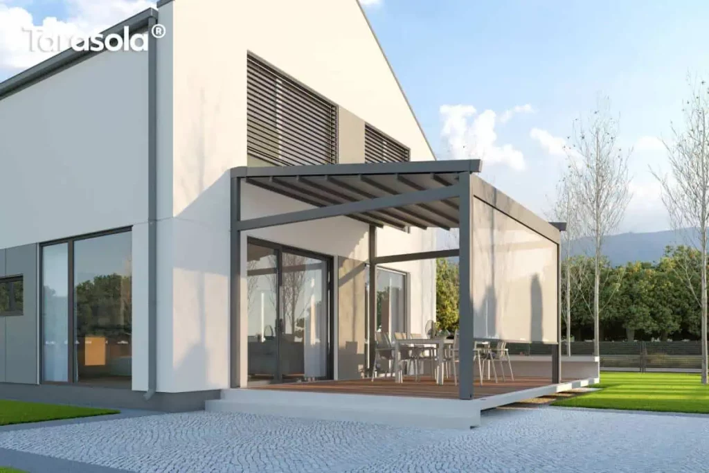 Nowoczesna pergola tarasowa Elegnacy Tarasola o aluminiowej konstrukcji w kolorze ciemnoszarym, przytwierdzona do białej elewacji piętrowego budynku mieszkalnego. Zadaszenie wykonane jest z ruchomej, segmentowej tkaniny w kolorze szarym, a na bocznej ścianie konstrukcji zamontowano pionową osłonę przeciwsłoneczną typu roleta ZIP screen, która jest częściowo opuszczona. Pod pergolą, na podwyższonym tarasie wyłożonym deskami kompozytowymi w kolorze naturalnego drewna, znajduje się strefa jadalniana z dużym stołem i krzesłami ogrodowymi. Budynek posiada nowoczesną architekturę z dużymi oknami tarasowymi oraz zewnętrznymi żaluzjami fasadowymi na piętrze. Teren przed tarasem wybrukowano jasną kostką granitową, a obok widoczny jest fragment zielonego trawnika oraz drzewa i pasmo górskie w oddali pod jasnym, słonecznym niebem.