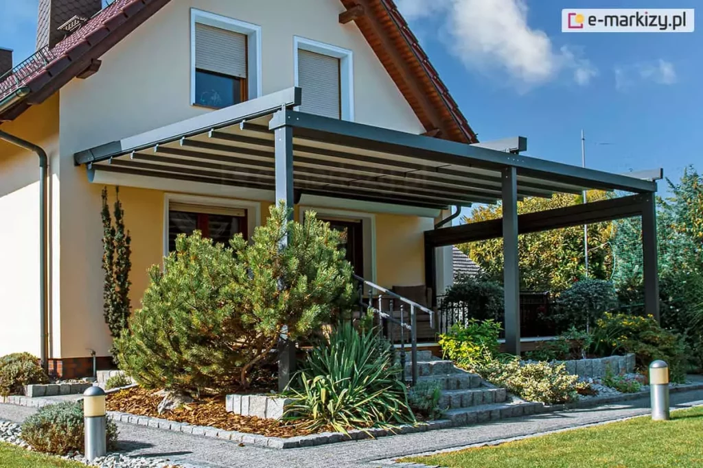 Nowoczesna pergola tarasowa przyścienna Solid Aluprof o konstrukcji aluminiowej w kolorze ciemnego antracytu, zamontowana do elewacji piętrowego domu jednorodzinnego o jasnych ścianach. Zadaszenie pergoli składa się z segmentowego, ruchomego poszycia tkaninowego w kolorze jasnoszarym, które wspiera się na poprzecznych profilach aluminiowych. Konstrukcja oparta jest na masywnych słupach frontowych, a boczna część tarasu została dodatkowo wyposażona w poziomą belkę wzmacniającą oraz roletę refleksol Ziiip. Taras znajduje się na podwyższeniu, na które prowadzą kamienne schody z metalową balustradą. Wokół tarasu zaprojektowano estetyczny ogród z dużą sosną górską, juką oraz innymi niskimi krzewami ozdobnymi na podłożu z kory sosnowej, oddzielonym kamiennymi obrzeżami od ścieżki z jasnej kostki brukowej. Wzdłuż ścieżki i trawnika ustawione są nowoczesne, metalowe lampy ogrodowe w kształcie słupków z mlecznymi kloszami. W tle widoczna jest bryła budynku z brązowym dachem oraz oknami wyposażonymi w zewnętrzne rolety okienne. Zdjęcie wykonane w słoneczny dzień przy błękitnym niebie, prezentuje harmonijne połączenie architektury ogrodowej z otoczeniem roślinnym.