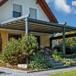 Nowoczesna pergola tarasowa przyścienna Solid Aluprof o konstrukcji aluminiowej w kolorze ciemnego antracytu, zamontowana do elewacji piętrowego domu jednorodzinnego o jasnych ścianach. Zadaszenie pergoli składa się z segmentowego, ruchomego poszycia tkaninowego w kolorze jasnoszarym, które wspiera się na poprzecznych profilach aluminiowych. Konstrukcja oparta jest na masywnych słupach frontowych, a boczna część tarasu została dodatkowo wyposażona w poziomą belkę wzmacniającą oraz roletę refleksol Ziiip. Taras znajduje się na podwyższeniu, na które prowadzą kamienne schody z metalową balustradą. Wokół tarasu zaprojektowano estetyczny ogród z dużą sosną górską, juką oraz innymi niskimi krzewami ozdobnymi na podłożu z kory sosnowej, oddzielonym kamiennymi obrzeżami od ścieżki z jasnej kostki brukowej. Wzdłuż ścieżki i trawnika ustawione są nowoczesne, metalowe lampy ogrodowe w kształcie słupków z mlecznymi kloszami. W tle widoczna jest bryła budynku z brązowym dachem oraz oknami wyposażonymi w zewnętrzne rolety okienne. Zdjęcie wykonane w słoneczny dzień przy błękitnym niebie, prezentuje harmonijne połączenie architektury ogrodowej z otoczeniem roślinnym.