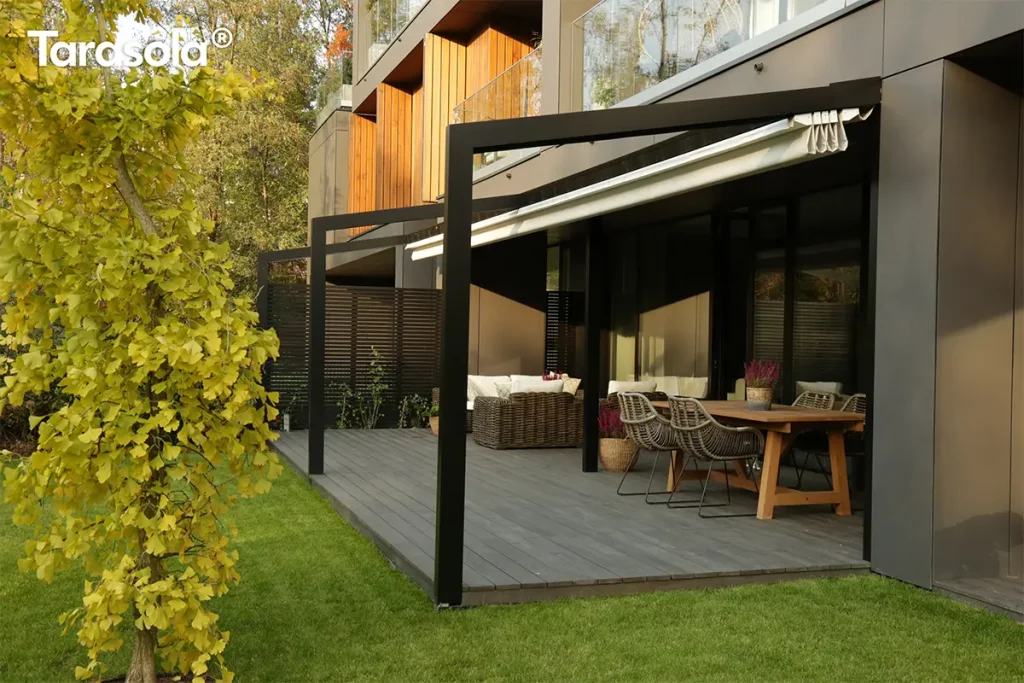 Czarna aluminiowa pergola tarasowa Tarasola Light o konstrukcji przyściennej, zamontowana na nowoczesnym tarasie wyłożonym szarymi deskami kompozytowymi. Stelaż pergoli składa się z minimalistycznych, ciemnych profili o kwadratowym przekroju, a dach tworzy jasna, kremowa tkanina techniczna, która na zdjęciu jest częściowo zwinięta w efektowne fałdy przy elewacji budynku. Pod zadaszeniem zaaranżowano strefę wypoczynkową z dużym drewnianym stołem, przy którym stoją plecione krzesła w kolorze szarym, oraz zestawem mebli technorattanowych z jasnymi poduszkami w głębi. Elewacja budynku jest nowoczesna, wykonana z ciemnych paneli z elementami naturalnego drewna i dużymi przeszkleniami. Po lewej stronie kompozycji, na pierwszym planie, widoczne jest ozdobne drzewo o intensywnie żółtych liściach, a w tle soczysty zielony trawnik oraz gęsta roślinność ogrodowa. Światło słoneczne pada pod kątem, tworząc wyraźne cienie konstrukcji na tarasie i podkreślając fakturę materiałów.