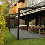 Czarna aluminiowa pergola tarasowa Tarasola Light o konstrukcji przyściennej, zamontowana na nowoczesnym tarasie wyłożonym szarymi deskami kompozytowymi. Stelaż pergoli składa się z minimalistycznych, ciemnych profili o kwadratowym przekroju, a dach tworzy jasna, kremowa tkanina techniczna, która na zdjęciu jest częściowo zwinięta w efektowne fałdy przy elewacji budynku. Pod zadaszeniem zaaranżowano strefę wypoczynkową z dużym drewnianym stołem, przy którym stoją plecione krzesła w kolorze szarym, oraz zestawem mebli technorattanowych z jasnymi poduszkami w głębi. Elewacja budynku jest nowoczesna, wykonana z ciemnych paneli z elementami naturalnego drewna i dużymi przeszkleniami. Po lewej stronie kompozycji, na pierwszym planie, widoczne jest ozdobne drzewo o intensywnie żółtych liściach, a w tle soczysty zielony trawnik oraz gęsta roślinność ogrodowa. Światło słoneczne pada pod kątem, tworząc wyraźne cienie konstrukcji na tarasie i podkreślając fakturę materiałów.