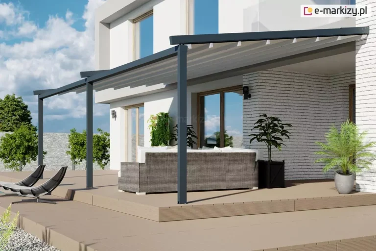 Nowoczesna pergola przyścienna o antracytowej konstrukcji aluminiowej, zamontowana nad wielopoziomowym tarasem wykonanym z jasnych desek kompozytowych. System opiera się na trzech pionowych słupach nośnych, które podtrzymują długą ramę z zadaszeniem z białej, odpornej na warunki atmosferyczne tkaniny, wspartej na poprzecznych profilach. Pod pergolą, przy ścianie z białej cegły dekoracyjnej, ustawiono duży narożnik ogrodowy z technorattanu w kolorze szarym, a obok niego znajdują się rośliny w czarnych i szarych donicach. Na niższym poziomie tarasu widoczne są dwa nowoczesne, ciemne leżaki o ergonomicznym kształcie. Całość aranżacji dopełnia minimalistyczna, biała elewacja budynku z dużymi przeszkleniami, w których odbija się błękitne niebo z delikatnymi chmurami.