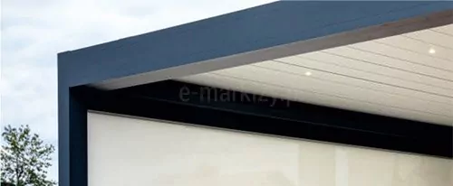Prezentowane ujęcie architektoniczne skupia się na zewnętrznym narożniku nowoczesnej pergoli tarasowej, model Tarasola Technic Climatic ukazując połączenie masywnej ramy konstrukcyjnej zintegrowanej z pionową roletą typu screen. W górnej części kadru widoczny jest solidny, antracytowy profil okapowy o geometrycznym, prostokątnym przekroju, który tworzy wyraźny, elegancki kontur zadaszenia na tle jasnego, zachmurzonego nieba. Pod główną belką nośną zamontowano segment sufitowy składający się z białych, równoległych lameli dachowych, w które wkomponowano punktowe oświetlenie LED emitujące ciepłe, rozproszone światło. W lewej części kompozycji pionowy słup narożny o tej samej ciemnoszatej barwie łączy się bezszwowo z poziomym wieńcem, tworząc stabilną oprawę dla zamontowanej w świetle konstrukcji osłony bocznej. W dolnej i środkowej partii kadru znajduje się rozwinięta roleta z półprzepuszczalnej tkaniny technicznej w jasnym, kremowobiałym odcieniu, zwinięta częściowo w górnej kaszcie ukrytej za czarnym profilem poprzecznym, co skutecznie chroni strefę wypoczynkową przed wiatrem i słońcem. W lewym dolnym rogu kadru widoczny jest fragment zieleni naturalnej w postaci liści drzewa, co osadza konstrukcję w realnym, przydomowym otoczeniu, a cała kompozycja charakteryzuje się precyzyjnym wykończeniem detali montażowych oraz nowoczesną estetyką architektoniczną.