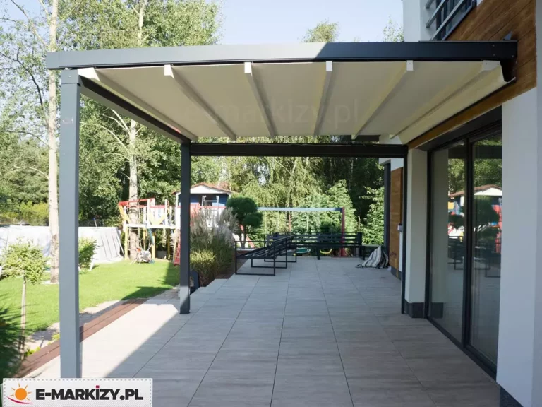 Nowoczesna pergola tarasowa Luxo na wymiar z ruchomym zadaszeniem tkaninowym, zamontowana do białej elewacji budynku z drewnianymi wstawkami. Konstrukcja o antracytowym kolorze opiera się na dwóch solidnych słupach nośnych, podtrzymujących rozłożone, jasne poszycie materiałowe wsparte na poprzecznych profilach aluminiowych. Pod zadaszeniem znajduje się przestronny taras wyłożony jasnymi, wielkoformatowymi płytami, na którym ustawiono minimalistyczne, czarne ławki ogrodowe. W tle widoczny jest ogród z trawnikiem, drzewami liściastymi oraz placem zabaw dla dzieci, co podkreśla funkcjonalność konstrukcji w przydomowej strefie rekreacyjnej. Całość kompozycji, uchwycona w dziennym świetle, prezentuje estetyczne połączenie nowoczesnej architektury z otaczającą naturą.