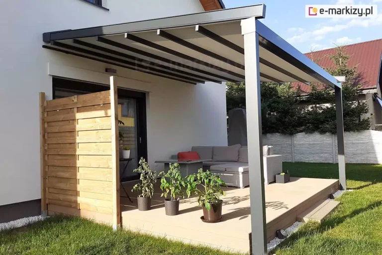 Przyścienna pergola tarasowa Square o antracytowej, aluminiowej konstrukcji, zamontowana przy białej elewacji domu jednorodzinnego. Zadaszenie pergoli wykonane jest z jasnego materiału rozpiętego na poprzecznych, ciemnych profilach, a jego tył przymocowano bezpośrednio do ściany budynku nad drzwiami tarasowymi. Konstrukcja wspiera się na dwóch solidnych, pionowych słupach o kwadratowym przekroju, które są osadzone w podłożu, tuż przy krawędzi tarasu. Na tarasie wyłożonym jasnymi deskami kompozytowymi urządzono strefę wypoczynkową z szarym narożnikiem technorattanowym i dopasowanym stolikiem kawowym. Po lewej stronie tarasu zamontowano pionową osłonę z jasnych, drewnianych desek, która pełni funkcję parawanu. W doniczkach przed narożnikiem rosną trzy krzewy pomidorów, a w tle widać biały, dekoracyjny płot betonowy oraz wysokie drzewa iglaste. Całość kompozycji dopełnia zadbany zielony trawnik otaczający taras, a scena oświetlona jest naturalnym światłem słonecznym.