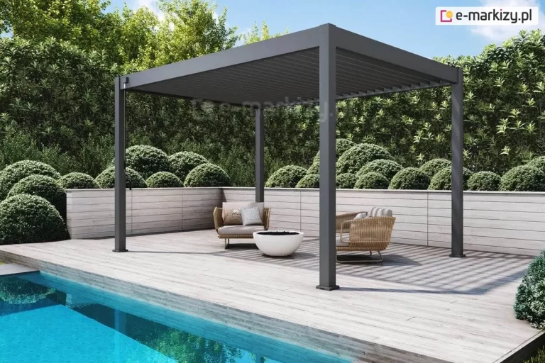 Nowoczesna pergola lamelowa wolnostojąca Marlo w kolorze antracytowym, zainstalowana na tarasie z jasnych desek drewnopodobnych tuż przy krawędzi basenu. Konstrukcja wspiera się na czterech narożnych słupach o kwadratowym przekroju, a jej dach tworzą ruchome lamele aluminiowe ustawione w pozycji zamkniętej. Pod zadaszeniem znajduje się minimalistyczna strefa wypoczynkowa z dwoma fotelami o beżowym splocie, wyposażonymi w jasne poduszki, oraz niskie palenisko ogrodowe w formie białej misy. Taras jest otoczony niskim murkiem z poziomych paneli w jasnym kolorze, za którym rozciąga się gęsty żywopłot z formowanych w kule krzewów liściastych. W lewym dolnym rogu widoczna jest krystalicznie czysta, błękitna woda basenu, w której odbijają się fragmenty architektury i roślinności. Przy jednym ze słupów pergoli zamontowany jest mechanizm korbowy służący do ręcznej regulacji kąta nachylenia lameli dachowych.