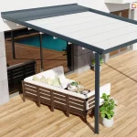 Widok z góry na nowoczesną pergolę Luxo o antracytowej konstrukcji, zamontowaną na przestronnym tarasie wyłożonym jasnymi deskami. Aluminiowa rama z zadaszeniem z białej, napiętej tkaniny chroni strefę wypoczynkową, w której ustawiono narożną sofę ogrodową z ciemnego drewna z białymi poduchami. Nad częścią materiału widoczny jest systemowy daszek ochronny z lamelami w kolorze konstrukcji, zabezpieczający poszycie po jego zwinięciu. W otoczeniu pergoli znajduje się basen, rower oparty o nowoczesną elewację budynku oraz donice z roślinami, co podkreśla rekreacyjny charakter przydomowej przestrzeni.
