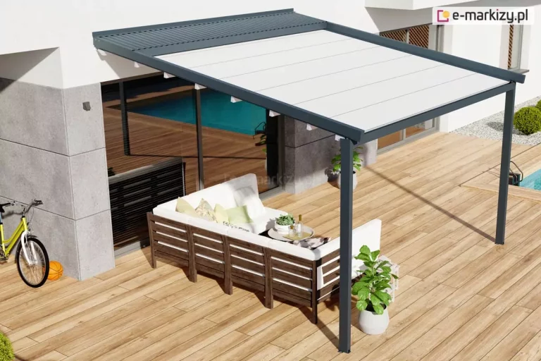 Widok z góry na nowoczesną pergolę Luxo o antracytowej konstrukcji, zamontowaną na przestronnym tarasie wyłożonym jasnymi deskami. Aluminiowa rama z zadaszeniem z białej, napiętej tkaniny chroni strefę wypoczynkową, w której ustawiono narożną sofę ogrodową z ciemnego drewna z białymi poduchami. Nad częścią materiału widoczny jest systemowy daszek ochronny z lamelami w kolorze konstrukcji, zabezpieczający poszycie po jego zwinięciu. W otoczeniu pergoli znajduje się basen, rower oparty o nowoczesną elewację budynku oraz donice z roślinami, co podkreśla rekreacyjny charakter przydomowej przestrzeni.