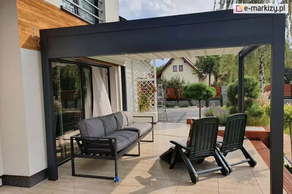 Zdjęcie prezentuje nowoczesną pergolę tarasową Luxury w ujęciu od boku, zamontowaną jako zadaszenie przyścienne na tarasie z jasnych płyt ceramicznych. Grafitowa konstrukcja z aluminium o matowym wykończeniu składa się z szerokiej ramy czołowej oraz bocznej, wspartej na pionowych słupach o kwadratowym przekroju. Pod dachem, wykonanym z jasnego, segmentowego poszycia materiałowego, znajduje się strefa wypoczynkowa wyposażona w szarą sofę o metalowej konstrukcji oraz dwa rozkładane fotele ogrodowe. W tle widoczna jest elewacja domu z białym tynkiem i wstawkami z naturalnego drewna, a także biała, ozdobna kratka ogrodowa z roślinnością pnącą. Za tarasem rozpościera się widok na sąsiedni budynek z dwuspadowym dachem oraz wysokie drzewa liściaste. Ujęcie w świetle dziennym podkreśla estetyczne połączenie ciemnej konstrukcji pergoli z jasnym zadaszeniem i elementami architektury ogrodu.