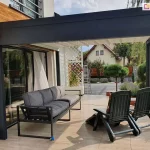 Zdjęcie prezentuje nowoczesną pergolę tarasową Luxury w ujęciu od boku, zamontowaną jako zadaszenie przyścienne na tarasie z jasnych płyt ceramicznych. Grafitowa konstrukcja z aluminium o matowym wykończeniu składa się z szerokiej ramy czołowej oraz bocznej, wspartej na pionowych słupach o kwadratowym przekroju. Pod dachem, wykonanym z jasnego, segmentowego poszycia materiałowego, znajduje się strefa wypoczynkowa wyposażona w szarą sofę o metalowej konstrukcji oraz dwa rozkładane fotele ogrodowe. W tle widoczna jest elewacja domu z białym tynkiem i wstawkami z naturalnego drewna, a także biała, ozdobna kratka ogrodowa z roślinnością pnącą. Za tarasem rozpościera się widok na sąsiedni budynek z dwuspadowym dachem oraz wysokie drzewa liściaste. Ujęcie w świetle dziennym podkreśla estetyczne połączenie ciemnej konstrukcji pergoli z jasnym zadaszeniem i elementami architektury ogrodu.