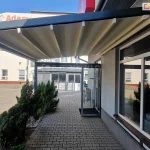 Zdjęcie prezentuje nowoczesną pergolę Square MOL przyścienną, zainstalowaną nad chodnikiem przy budynku usługowym lub biurowym. Konstrukcja pergoli charakteryzuje się minimalistycznym designem i wykonana jest z profili aluminiowych o kwadratowym przekroju w kolorze ciemnego grafitu lub antracytu. Zadaszenie stanowi ruchoma, segmentowa tkanina w kolorze kremowym lub jasnobeżowym, która jest zamocowana na prowadnicach i tworzy płaską płaszczyznę pod kątem nachylenia umożliwiającym odpływ wody. Pergola Square MOL przyścienna wsparta jest z jednej strony na elewacji budynku, a z drugiej na solidnym pionowym słupie, co tworzy funkcjonalne zadaszenie nad wejściem do obiektu i wzdłuż dużych witryn okiennych. Podłoże pod konstrukcją wyłożone jest szarą kostką brukową, a wzdłuż krawędzi chodnika rosną niskie, gęste iglaki. W tle widoczny jest plac wyłożony kostką oraz biały budynek z czerwonymi akcentami przy fundamencie i widocznym napisem na elewacji. Zdjęcie zostało wykonane w słoneczny dzień.