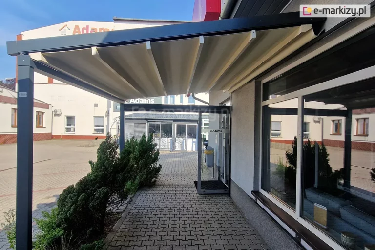 Zdjęcie prezentuje nowoczesną pergolę Square MOL przyścienną, zainstalowaną nad chodnikiem przy budynku usługowym lub biurowym. Konstrukcja pergoli charakteryzuje się minimalistycznym designem i wykonana jest z profili aluminiowych o kwadratowym przekroju w kolorze ciemnego grafitu lub antracytu. Zadaszenie stanowi ruchoma, segmentowa tkanina w kolorze kremowym lub jasnobeżowym, która jest zamocowana na prowadnicach i tworzy płaską płaszczyznę pod kątem nachylenia umożliwiającym odpływ wody. Pergola Square MOL przyścienna wsparta jest z jednej strony na elewacji budynku, a z drugiej na solidnym pionowym słupie, co tworzy funkcjonalne zadaszenie nad wejściem do obiektu i wzdłuż dużych witryn okiennych. Podłoże pod konstrukcją wyłożone jest szarą kostką brukową, a wzdłuż krawędzi chodnika rosną niskie, gęste iglaki. W tle widoczny jest plac wyłożony kostką oraz biały budynek z czerwonymi akcentami przy fundamencie i widocznym napisem na elewacji. Zdjęcie zostało wykonane w słoneczny dzień.