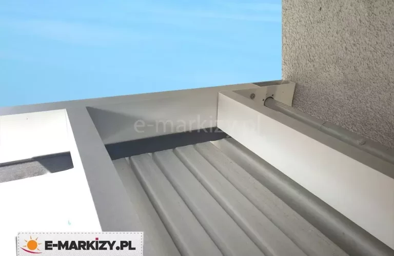 Techniczne zbliżenie z perspektywy górnej na system montażu białej pergoli tarasowej do elewacji budynku o chropowatej strukturze. Zdjęcie szczegółowo ukazuje profil przyścienny oraz mechanizm prowadnic, w których osadzone jest złożone zadaszenie tkaninowe ułożone w gęste, regularne plisy. Widoczny jest aluminiowy profil poprzeczny pełniący funkcję osłony oraz stalowy wał napędowy umieszczony w bliskiej odległości od ściany, co zapewnia stabilność i płynność pracy systemu rozsuwania. Całość konstrukcji jest polakierowana na biały kolor o matowym wykończeniu, a precyzyjne łączenia elementów podkreślają wysoką jakość wykonania detali architektonicznych. W tle, ponad krawędzią ramy, widoczna jest jednolita tafla błękitnego nieba, co pozwala wyeksponować geometrię i czystość linii profilu aluminiowego.