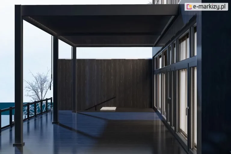 Antracytowa pergola bioklimatyczna Marlo w wersji przyściennej, zamontowana nad tarasem nowoczesnego budynku. Konstrukcja zadaszenia wspiera się na trzech pionowych, kwadratowych słupach od lewej, a po prawej stronie jest przytwierdzona bezpośrednio do elewacji obiektu charakteryzującego się dużą powierzchnią przeszkleń. Lamele dachowe są ustawione w pozycji zamkniętej, tworząc szczelną, płaską osłonę nad powierzchnią tarasu. Podłoże tarasu wykonane jest z ciemnych desek o delikatnym połysku, na których widać wyraźny cień rzucany przez dach pergoli. Po lewej stronie kadr domyka czarna balustrada o prostej formie, za którą widoczna jest zimowa sceneria z ośnieżonym terenem i bezlistnym drzewem pokrytym szronem. Perspektywa zdjęcia z poziomu tarasu pozwala dostrzec wysokość konstrukcji oraz sposób jej integracji z architekturą domu i otaczającym krajobrazem.
