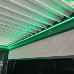 Zdjęcie prezentuje szczegółowe ujęcie wewnętrznej konstrukcji nowoczesnej pergoli tarasowej z dachem lamelowym, wykonane z perspektywy dolnej. Głównym elementem zadaszenia są białe, aluminiowe lamele ułożone równolegle obok siebie, które na krawędziach posiadają charakterystyczne wyprofilowanie ułatwiające odprowadzanie wody. Wzdłuż wewnętrznej ramy konstrukcyjnej, na styku z dachem, zamontowano liniowe oświetlenie LED, które emituje intensywne światło o nasyconej, zielonej barwie. Zielona poświata oświetla krawędzie lameli oraz górną część bocznej osłony. Poniżej linii światła widoczna jest pionowa, jasna roleta typu screen, która jest częściowo rozwinięta, a jej powierzchnia delikatnie odbija zielony blask. Rama pergoli ma ciemny, prawdopodobnie antracytowy kolor, który kontrastuje z jasnymi lamelami i jaskrawym oświetleniem. W lewym dolnym rogu zdjęcia, przez przeszklony fragment konstrukcji, prześwituje zarysy wnętrza pomieszczenia lub hali wystawowej, sugerując, że zdjęcie mogło zostać wykonane w salonie ekspozycyjnym.