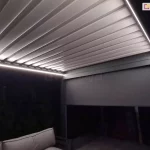 Zdjęcie przedstawia nowoczesną pergolę tarasową wyposażoną w dach lamelowy oraz oświetlenie LED, sfotografowaną w warunkach nocnych lub przy bardzo słabym oświetleniu zewnętrznym. Konstrukcja zadaszenia składa się z szeregu równolegle ułożonych, płaskich lameli w jasnym kolorze, które tworzą szczelną płaszczyznę dachu. Wzdłuż wewnętrznych krawędzi ramy pergoli zamontowano liniowe paski LED, które emitują intensywne, chłodne białe światło, oświetlając spodnią stronę lameli oraz przestrzeń pod konstrukcją. Po prawej stronie widoczny jest pionowy słupek nośny pergoli oraz zamontowana boczna roleta typu screen w ciemnym odcieniu, która jest częściowo rozwinięta, zasłaniając górną partię boku konstrukcji. W dolnej części kadru, po lewej stronie, widoczny jest fragment mebla ogrodowego, prawdopodobnie sofy, z jasnymi poduszkami oparciowymi. Tło za pergolą jest ciemne i niewyraźne, sugerując obecność roślinności ogrodowej w nocy. Całość kompozycji skupia się na detalach technicznych systemu oświetlenia oraz strukturze domkniętego dachu lamelowego.