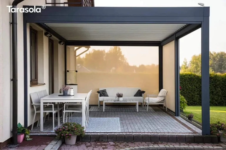 Nowoczesna aluminiowa pergola lamelowa przyścienna Tarasola Essential w eleganckim, ciemnoantracytowym kolorze, zamontowana jako konstrukcja przyścienna do elewacji budynku. Model Essential Tarasola charakteryzuje się płaskim dachem z ruchomymi lamelami, które na zdjęciu są ustawione w pozycji zamkniętej, tworząc szczelną barierę chroniącą przed słońcem i deszczem. Konstrukcja wspiera się na solidnych, kwadratowych słupach bocznych, które precyzyjnie wyznaczają strefę wypoczynku. Lewa strona oraz tył pergoli zostały wyposażone w jasne przesłony przeciwsłoneczne typu screen, które przepuszczają miękkie, rozproszone światło, jednocześnie odgradzając wnętrze tarasu od otoczenia i wiatru. Pod zadaszeniem Essential Tarasola znajduje się starannie zaaranżowana przestrzeń podzielona na część jadalnianą i salonową, ułożona na szarej kostce brukowej z ozdobnym obramowaniem z ciemniejszej kostki. Po lewej stronie ustawiono biały stół ogrodowy z czterema dopasowanymi kolorystycznie krzesłami o lekkiej konstrukcji, umieszczony na niebiesko-białym dywanie zewnętrznym w geometryczne wzory. W głębi tarasu, pod tylną przesłoną screen, znajduje się strefa relaksu z białą sofą ogrodową udekorowaną ciemnymi poduszkami, niskim stolikiem kawowym oraz pojedynczym fotelem z jasnym narzutem, również ustawionymi na ozdobnym dywanie. Na jasnej elewacji domu widoczne są brązowe ramy okienne, czarna rura spustowa oraz zamontowane pod dachem pergoli oświetlenie zewnętrzne w formie punktowych reflektorów. Na pierwszym planie, przy krawędzi tarasu wykończonego ciemną palisadą, stoi donica z kwitnącymi różowymi kwiatami, a po prawej stronie rozciąga się zielony ogród z przystrzyżonym żywopłotem i trawnikiem. Całość aranżacji podkreśla nowoczesny design produktu Essential Tarasola, łącząc funkcjonalność z minimalistyczną estetyką.