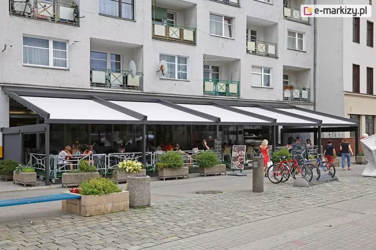 Wielomodułowa, wielkoformatowa pergola tarasowa Solid Aluprof o konstrukcji aluminiowej w kolorze antracytowym, zainstalowana jako zadaszenie ogródka restauracyjnego przy parterze wielokondygnacyjnego budynku mieszkalnego. System zadaszenia składa się z kilku sekcji białego poszycia tkaninowego, które jest w całości rozłożone pod kątem, zapewniając ochronę przed słońcem i opadami. Boczne ściany ogródka są częściowo oslonięte, tworząc zamkniętą przestrzeń dla gości. Wewnątrz ogródka widoczne są liczne stoliki z siedzącymi przy nich ludźmi oraz dekoracyjne barierki ochronne w kolorze białym. Przed restauracją na brukowanym chodniku ustawiono betonowe donice z zieloną roślinnością i kwiatami, stojaki na rowery z zaparkowanymi jednośladami oraz tablice reklamowe typu potykacz. W tle widoczna jest jasna elewacja bloku z licznymi oknami i balkonami, a po prawej stronie fragment miejskiego deptaka z przechodniami. Zdjęcie wykonane w świetle dziennym, prezentuje komercyjne zastosowanie nowoczesnych systemów zadaszeń w przestrzeni miejskiej.