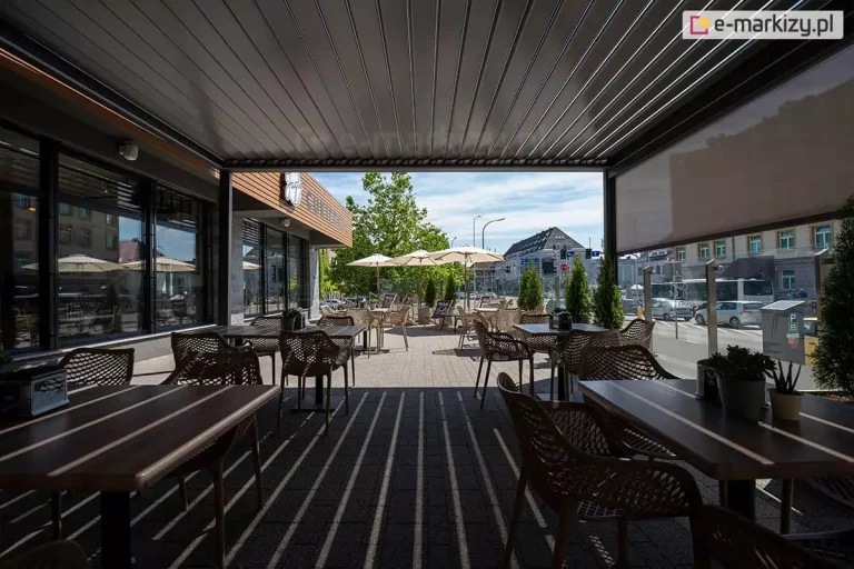 Widok z wnętrza zacienionej pergoli restauracyjnej na słoneczny taras zewnętrzny i fragment miasta. Konstrukcja posiada zadaszenie z ciemnych, aluminiowych lameli, które rzucają liniowy cień na podłoże z kostki brukowej. W strefie pod zadaszeniem oraz na otwartym tarasie ustawione są stoliki o drewnianych blatach z nowoczesnymi, ażurowymi krzesłami w kolorze beżowym. Po prawej stronie widoczna jest opuszczona do połowy przesłona boczna typu screen. W tle znajdują się białe parasole ogrodowe, donice z zielonymi tujami oraz budynek z szyldem restauracji. W oddali widoczna jest ulica z poruszającymi się samochodami i miejska zabudowa. Zdjęcie prezentuje praktyczne wykorzystanie pergoli bioklimatycznej do wydzielenia komfortowej strefy gastronomicznej o zróżnicowanym stopniu nasłonecznienia.