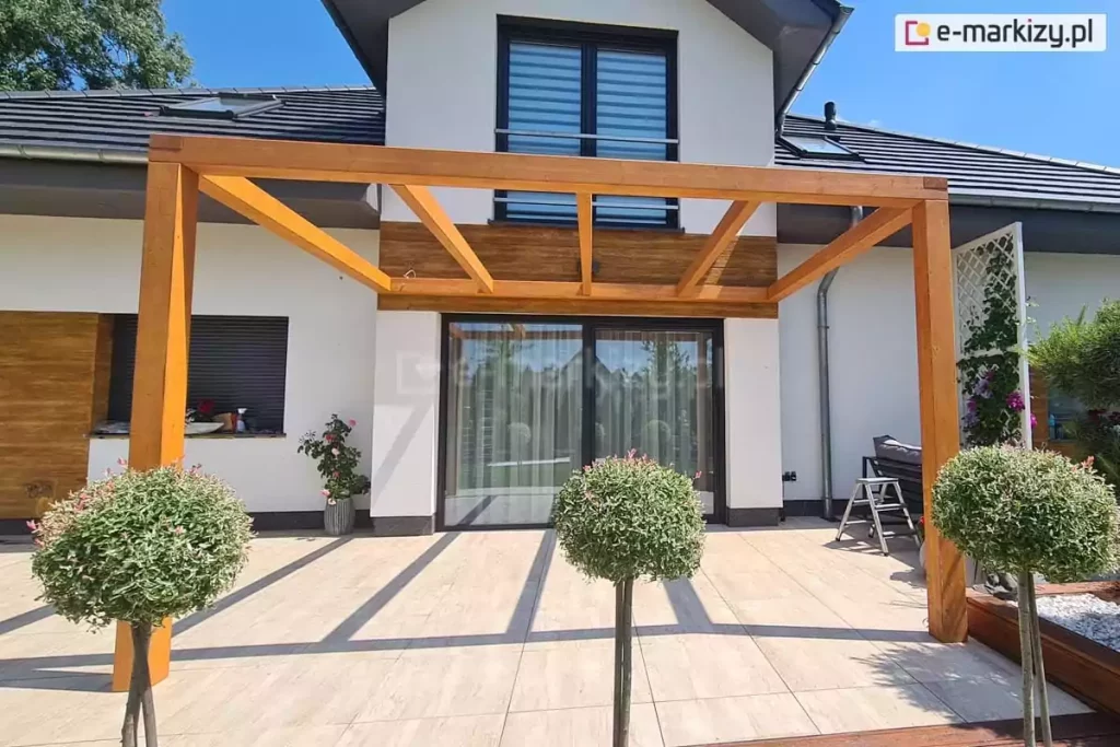 Pergola drewniana Juko przyścienna pokryta lakierobejcą z trzema wewnętrznymi krokwiami ustawiona na tarasie przy domu o białej elewacji z dużym oknem tarasowym o antracytowej ramie. Na pierwszym planie trzy niskie drzewka o kulisto przystrzyżonych koronach na cienkich pniach.