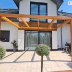 Pergola drewniana Juko przyścienna pokryta lakierobejcą z trzema wewnętrznymi krokwiami ustawiona na tarasie przy domu o białej elewacji z dużym oknem tarasowym o antracytowej ramie. Na pierwszym planie trzy niskie drzewka o kulisto przystrzyżonych koronach na cienkich pniach.