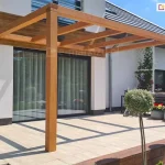 Pergola drewniana przyścienna Juko stanowiąca częściowe zadaszenie tarasu z jasnej płytki gresowej, na którym ustawiono wyłącznie doniczkę z kwiatami. W otoczeniu tarasu widoczne liczne rośliny.