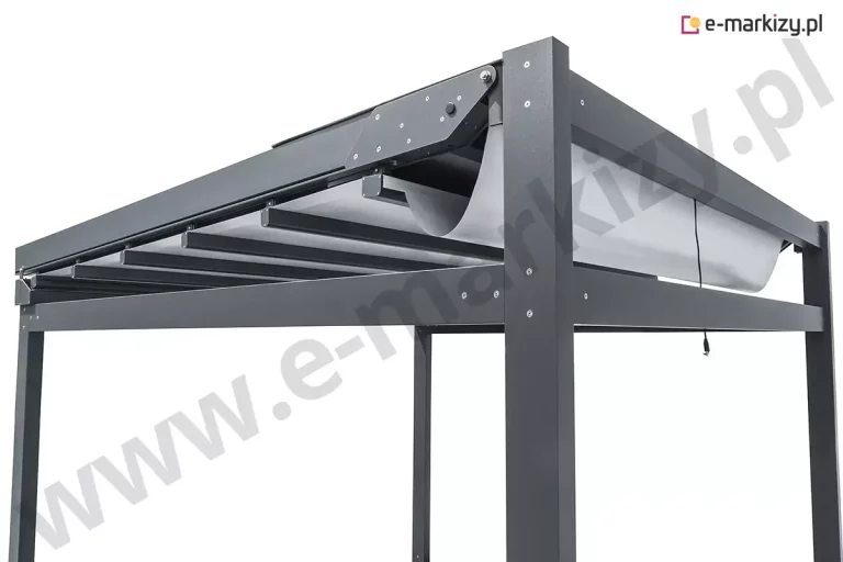 Szczegółowe ujęcie górnego węzła konstrukcyjnego pergoli tarasowej Solid Aluprof prezentujące mechanizm prowadzenia tkaniny oraz połączenia profili aluminiowych w kolorze antracytowym. Na zbliżeniu widoczny jest masywny narożnik konstrukcji, gdzie pionowy słup łączy się z poziomą belką obwodową oraz szyną jezdną zadaszenia. System zadaszenia składa się z jasnego, technicznego materiału tekstylnego, który jest rozpięty na poprzecznych profilach przeciwwietrznych poruszających się wewnątrz bocznych prowadnic. Materiał w tylnej części tworzy estetyczną, swobodnie zwisającą falę, co jest charakterystyczne dla systemów rzymskich przy niepełnym rozłożeniu. Detal ukazuje precyzyjne wykończenie elementów metalowych, w tym bocznych osłon mechanizmu napędowego o trójkątnym kształcie montowanych na śruby imbusowe oraz czarny przewód elektryczny wyprowadzony z ramy, służący do zasilania automatyki pogodowej lub silnika. Powierzchnia aluminium posiada delikatną strukturę lakieru proszkowego, a cała konstrukcja cechuje się wysoką sztywnością i nowoczesnym, surowym designem zorientowanym na funkcjonalność.