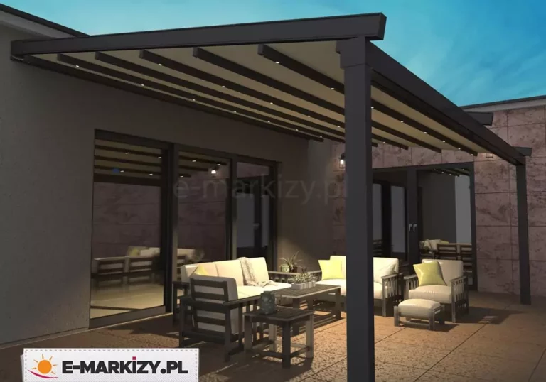 Nowoczesna pergola tarasowa o grafitowej, aluminiowej konstrukcji, zamontowana bezpośrednio do elewacji budynku o ciemnoszarym tynku. Zadaszenie pergoli wykonane jest z beżowego, segmentowego materiału, który został rozpięty na poziomych profilach wyposażonych w zintegrowane punktowe oświetlenie LED. Światło z listew dachowych rzuca ciepły blask na przestrzeń wypoczynkową znajdującą się poniżej. Pod pergolą ustawiony jest zestaw mebli ogrodowych składający się z dwóch jasnych sof, fotela z podnóżkiem oraz dwóch ciemnych stolików kawowych o różnej wysokości. Meble posiadają białe poduchy siedziskowe i dekoracyjne jasnozielone poduszki. W tle widoczne są duże przeszklenia drzwi tarasowych, w których odbija się oświetlona konstrukcja zadaszenia oraz fragmenty elewacji wyłożonej płytami o strukturze kamienia. Posadzka tarasu wykonana jest z dużych płyt w odcieniu przygaszonego brązu. Całość ujęcia prezentuje aranżację w porze wieczornej, podkreślając funkcjonalność oświetlenia i przytulną atmosferę miejsca.