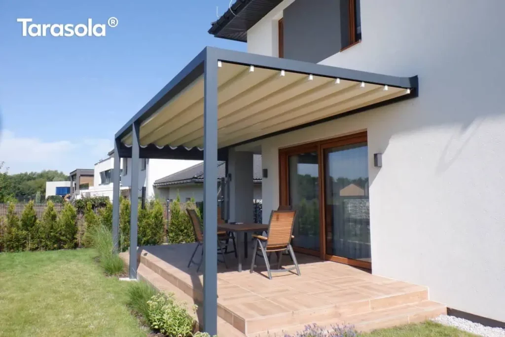 Elektryczna pergola tarasowa przyścienna Tarasola Flat o konstrukcji aluminiowej w kolorze antracytowym, zamontowana do białej elewacji piętrowego budynku. Zadaszenie stanowi jasny, kremowy dach tkaninowy, który jest w pełni rozwinięty, tworząc niemal płaską powierzchnię z delikatnymi, regularnymi liniami profili przeciwwietrznych. Konstrukcja wspiera się na dwóch solidnych, przednich słupach o kwadratowym przekroju, osłaniając taras wyłożony jasnobrązowymi płytkami ceramicznymi imitującymi drewno. W centralnej części tarasu ustawiono drewniany stół z czterema pasującymi krzesłami o konstrukcji metalowej z drewnianym wykończeniem oparć i siedzisk. Duże, przesuwne drzwi tarasowe w brązowych ramach zapewniają bezpośrednie wyjście z wnętrza domu pod zadaszenie. Otoczenie stanowi zadbany trawnik, niskie nasadzenia z traw ozdobnych i lawendy przy krawędzi tarasu oraz rząd tui tworzący żywopłot wzdłuż ogrodzenia. W tle widoczna jest nowoczesna zabudowa jednorodzinna pod czystym, błękitnym niebem.