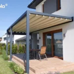 Elektryczna pergola tarasowa przyścienna Tarasola Flat o konstrukcji aluminiowej w kolorze antracytowym, zamontowana do białej elewacji piętrowego budynku. Zadaszenie stanowi jasny, kremowy dach tkaninowy, który jest w pełni rozwinięty, tworząc niemal płaską powierzchnię z delikatnymi, regularnymi liniami profili przeciwwietrznych. Konstrukcja wspiera się na dwóch solidnych, przednich słupach o kwadratowym przekroju, osłaniając taras wyłożony jasnobrązowymi płytkami ceramicznymi imitującymi drewno. W centralnej części tarasu ustawiono drewniany stół z czterema pasującymi krzesłami o konstrukcji metalowej z drewnianym wykończeniem oparć i siedzisk. Duże, przesuwne drzwi tarasowe w brązowych ramach zapewniają bezpośrednie wyjście z wnętrza domu pod zadaszenie. Otoczenie stanowi zadbany trawnik, niskie nasadzenia z traw ozdobnych i lawendy przy krawędzi tarasu oraz rząd tui tworzący żywopłot wzdłuż ogrodzenia. W tle widoczna jest nowoczesna zabudowa jednorodzinna pod czystym, błękitnym niebem.