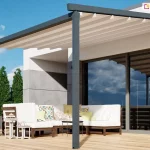 Nowoczesna pergola elektryczna Luxo zamontowana nad przestronnym tarasem z jasnych desek kompozytowych. Konstrukcja o antracytowym kolorze składa się z dwóch solidnych słupów nośnych oraz ramy zamocowanej do białej elewacji budynku, charakteryzującej się minimalistyczną architekturą i dużymi przeszkleniami. Zadaszenie wykonane z jasnej, kremowej tkaniny jest w pełni rozłożone i wsparte na poprzecznych profilach aluminiowych, tworząc geometryczny wzór na tle błękitnego nieba. Pod pergolą urządzono strefę wypoczynkową z dużym białym narożnikiem ogrodowym ozdobionym kolorowymi poduszkami, przy którym stoją niskie stoliki kawowe oraz ozdobne białe latarnie. W tle widoczne są starannie przycięte wysokie krzewy ogrodowe oraz fragment szklanych drzwi przesuwnych, w których odbijają się chmury i elementy tarasu.