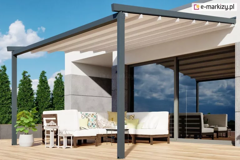 Nowoczesna pergola elektryczna Luxo zamontowana nad przestronnym tarasem z jasnych desek kompozytowych. Konstrukcja o antracytowym kolorze składa się z dwóch solidnych słupów nośnych oraz ramy zamocowanej do białej elewacji budynku, charakteryzującej się minimalistyczną architekturą i dużymi przeszkleniami. Zadaszenie wykonane z jasnej, kremowej tkaniny jest w pełni rozłożone i wsparte na poprzecznych profilach aluminiowych, tworząc geometryczny wzór na tle błękitnego nieba. Pod pergolą urządzono strefę wypoczynkową z dużym białym narożnikiem ogrodowym ozdobionym kolorowymi poduszkami, przy którym stoją niskie stoliki kawowe oraz ozdobne białe latarnie. W tle widoczne są starannie przycięte wysokie krzewy ogrodowe oraz fragment szklanych drzwi przesuwnych, w których odbijają się chmury i elementy tarasu.