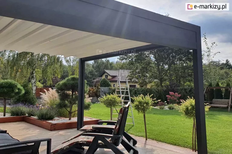 Zdjęcie prezentuje widok spod nowoczesnej pergoli tarasowej Luxury na zagospodarowany ogród w słoneczny dzień. Konstrukcja pergoli wykonana jest z matowych, grafitowych profili aluminiowych, a jej dach stanowi jasne, segmentowe poszycie materiałowe, które w tej chwili jest całkowicie rozłożone. Na pierwszym planie, w cieniu zadaszenia, widoczne są fragmenty mebli ogrodowych, w tym ciemne fotele z wzorzystymi poduszkami. Taras z jasnych płyt sąsiaduje bezpośrednio z zadbanym trawnikiem oraz licznymi nasadzeniami: niskimi drzewkami na pniu, ozdobnymi trawami i krzewami w drewnianych donicach wypełnionych białym kamieniem. W dalszej części ogrodu widać drewnianą huśtawkę, dom jednorodzinny z brązowym dachem oraz gęstą ścianę zieleni z drzew liściastych. Perspektywa zdjęcia podkreśla dużą przestrzeń wypoczynkową oraz skuteczną ochronę przed słońcem, jaką zapewnia rozpięty materiał pergoli.