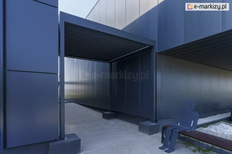 Przejście między budynkami osłonięte antracytową pergolą lamelową SB400 Aluprof z zamkniętym zadaszeniem z piór aluminiowych. W pobliżu przejścia ustawiono ławeczkę drewnianą z siedzącym na niej płaskim ludzikiem z aluminium w kolorze antracytowym.