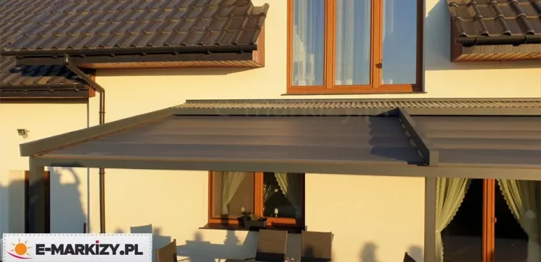 Zbliżenie na górną część konstrukcji antracytowej pergoli tarasowej Luxo w momencie pełnego rozłożenia zadaszenia tkaninowego. Zdjęcie wykonane z perspektywy frontowej ukazuje ciemnoszary profil prowadnicy bocznej zintegrowany z systemem uszczelnień oraz górny daszek ochronny wykonany z profilowanej blachy w tym samym kolorze co stelaż. Pod daszkiem widoczne jest napięte poszycie w ciemnym kolorze, które tworzy jednolitą, gładką płaszczyznę chroniącą taras przed opadami i słońcem. W tle widoczny jest fragment jasnej elewacji budynku, okno z brązową ramą oraz ciemna dachówka ceramiczna z systemem rynnowym. Oświetlenie naturalne podkreśla matowe wykończenie elementów aluminiowych oraz precyzyjne dopasowanie komponentów osłonowych dachu rzymskiego.