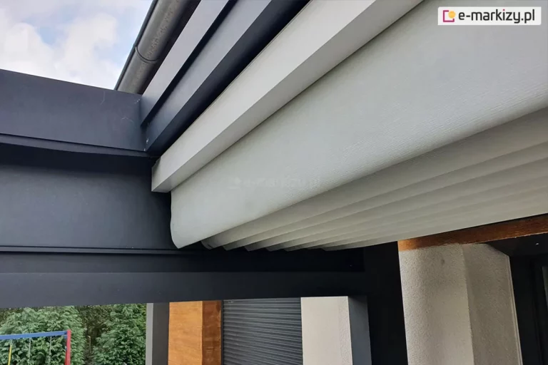 Zdjęcie przedstawia zbliżenie na techniczne detale wykończenia pergoli tarasowej Luxury w miejscu styku ruchomego zadaszenia z konstrukcją nośną. Na pierwszym planie widoczny jest masywny, poziomy profil aluminiowy w kolorze ciemnografitowym o matowej strukturze, który stanowi ramę systemu. Powyżej zamontowane jest jasnoszare, segmentowe poszycie wykonane z grubego materiału technicznego o wyraźnej teksturze, które jest w tej chwili częściowo złożone. Materiał układa się w estetyczne, równoległe fałdy podtrzymywane przez poprzeczne profile usztywniające w kolorze białym. W górnej części kadru widoczny jest fragment rynny oraz krawędź dachu budynku, co wskazuje na przyścienny sposób montażu konstrukcji. W tle, pod konstrukcją pergoli, dostrzec można fragment elewacji domu z elementami drewna, ciemną roletę okienną oraz zielone korony drzew ogrodowych. Ujęcie podkreśla precyzję dopasowania elementów oraz wysoką jakość materiałów użytych do produkcji zadaszenia.