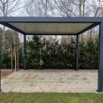 Widok od boku na wolnostojącą pergolę bioklimatyczną Sb350 Selt o antracytowej konstrukcji aluminiowej, ustawioną na tarasie z jasnej kostki brukowej. Pergola posiada cztery kwadratowe słupy nośne oraz dach z zamkniętymi lamelami w kolorze białym, tworzącymi szczelne zadaszenie. Konstrukcja jest zamontowana w ogrodzie; bezpośrednio za nią znajduje się gęsty pas zieleni składający się z wysokich tui, sosen oraz młodych brzóz wspartych na tyczkach. Po prawej stronie widoczny jest pień dużego drzewa liściastego oraz fragment czarnej donicy. Zdjęcie prezentuje symetryczną bryłę pergoli na tle naturalnej roślinności w pochmurny dzień.