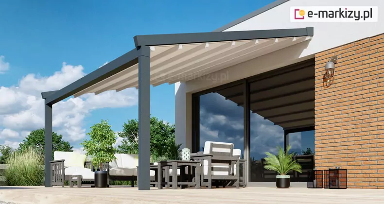 Zdjęcie przedstawia nowoczesną aranżację tarasu, na którym zamontowana została pergola przyścienna Moderno w kolorze ciemnoantracytowym. Konstrukcja wspiera się na solidnych, kwadratowych profilach aluminiowych zakotwiczonych w jasnym podłożu tarasu wykonanym z desek. Zadaszenie pergoli tworzy jasna, kremowa tkanina techniczna ułożona w charakterystyczne, równoległe fałdy, co wskazuje na segmentowy system składania poszycia. Materiał jest zamocowany na prowadnicach bocznych i wysunięty, osłaniając strefę wypoczynkową przed słońcem. Pergola jest przymocowana bezpośrednio do białej elewacji nowoczesnego budynku, tuż nad dużym, przesuwnym przeszkleniem tarasowym, w którym odbija się ogród. Część ściany budynku wykończona jest dekoracyjną cegłą w kolorze miodowym, na której zamontowano stylowy kinkiet zewnętrzny w formie metalowej klatki. Pod pergolą znajduje się zestaw mebli ogrodowych składający się z foteli i sof z białymi poduchami oraz ciemnych stolików kawowych. Przestrzeń jest ozdobiona roślinnością w donicach, w tym niską palmą oraz ozdobnymi trawami. W tle widoczne są zielone korony drzew oraz błękitne niebo z rozproszonymi, białymi chmurami, co podkreśla letni charakter prezentowanej pergoli przyściennej Moderno.