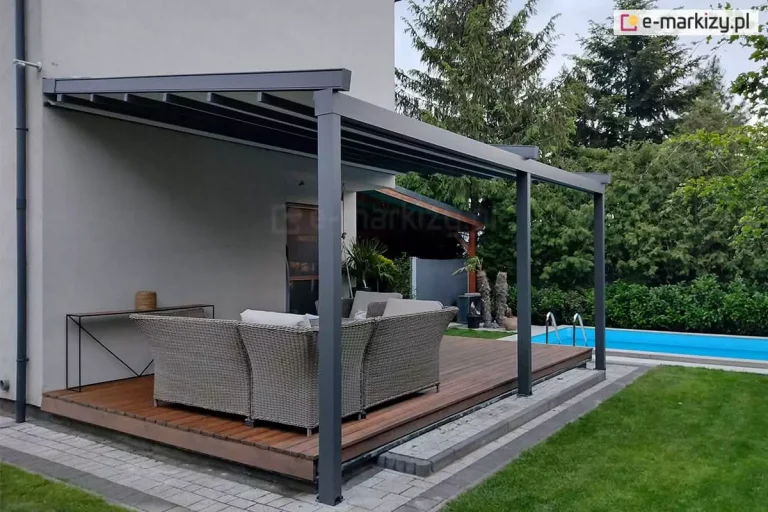 Zdjęcie przedstawia nowoczesną pergolę tarasową Square o konstrukcji aluminiowej w kolorze ciemnoszarym, antracytowym, zamontowaną przy elewacji piętrowego budynku mieszkalnego. Konstrukcja pergoli opiera się na dwóch solidnych pionowych słupach o kwadratowym przekroju, które są zakotwiczone w podłożu przy krawędzi tarasu. Zadaszenie pergoli jest płaskie, lekko nachylone w stronę ogrodu, z widocznymi poprzecznymi profilami wspierającymi poszycie. Pod pergolą znajduje się przestronna strefa wypoczynkowa urządzona na drewnianym tarasie wykonanym z desek o naturalnym, brązowym odcieniu. Wyposażenie strefy obejmuje duży, narożny zestaw mebli ogrodowych wykonanych z technorattanu w kolorze szarym, wyłożonych jasnymi, grubymi poduszkami na siedziskach i oparciach. Po lewej stronie, przy ścianie budynku, ustawiona jest długa, wąska konsola o minimalistycznym czarnym stelażu, na której stoi dekoracyjny element przypominający donicę lub lampion. Taras jest podwyższony względem reszty terenu i wykończony szarą kostką brukową tworzącą obramowanie. W tle po prawej stronie widoczny jest fragment niebieskiego basenu wpuszczonego w ziemię z metalową drabinką wejściową. Otoczenie stanowi zadbany ogród z zielonym trawnikiem oraz gęstym szpalerem wysokich drzew iglastych i krzewów, które tworzą naturalną barierę prywatności. Ściana domu, do której przylega pergola, jest otynkowana na jasnoszary kolor, a po lewej stronie widoczna jest pionowa rura spustowa systemu rynnowego w kolorze zbliżonym do ramy pergoli.