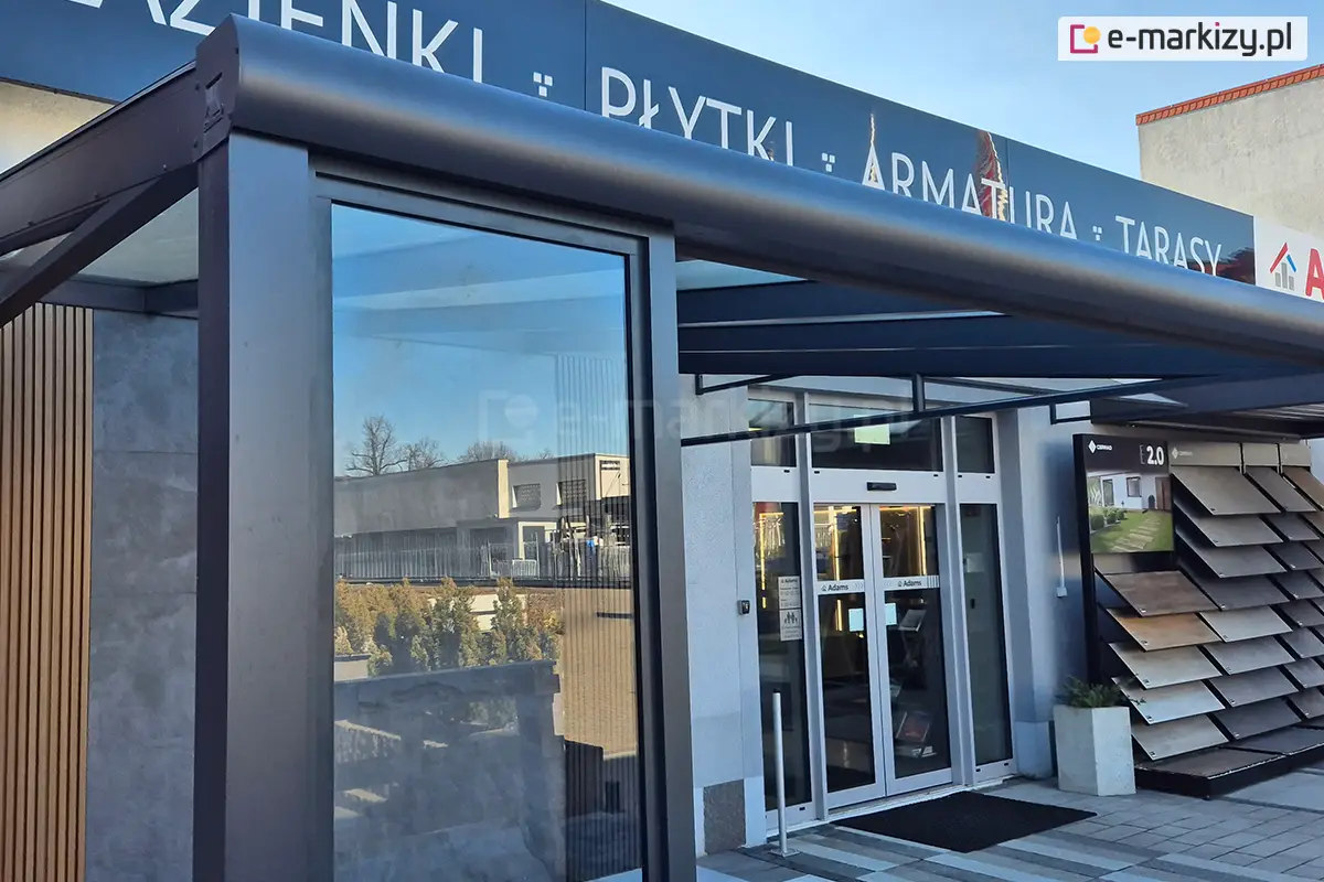 Ekspozycja zewnętrzna nowoczesnej pergoli tarasowej o konstrukcji aluminiowej w kolorze ciemnografitowym. Kluczowym elementem widocznym na pierwszym planie jest stałe przeszklenie boczne, oprawione w gruby profil pionowy, które pełni funkcję osłony przed wiatrem lub ścianki działowej. Nad ścianką stałą widoczna jest rynna zadaszenia o zaokrąglonym kształcie, a po lewej stronie widoczny jest fragment ściany wykończonej pionowymi lamelami drewnianymi oraz szarym spiekiem kwarcowym. W tle znajduje się wejście do salonu sprzedaży z przeszklonymi drzwiami automatycznymi oraz ekspozytor z próbkami materiałów elewacyjnych lub tarasowych. Całość ukazana jest w świetle dziennym, z wyraźnym odbiciem otoczenia i nieba w szybie stałego okna.