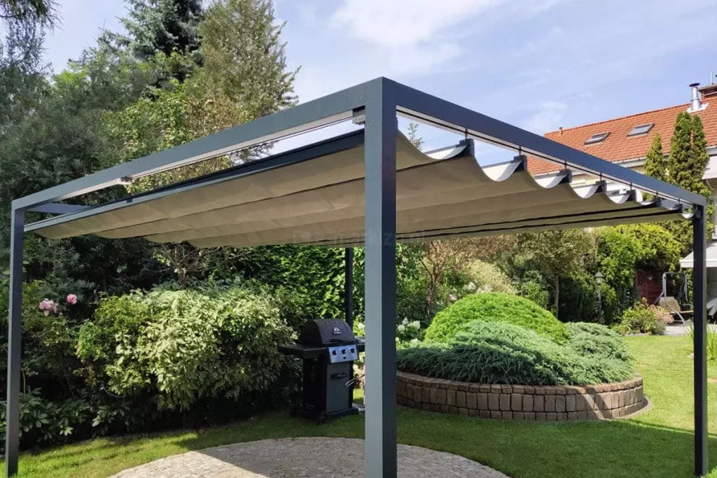 Wolnostojąca pergola rzymska Dragon o antracytowej, aluminiowej konstrukcji, umiejscowiona w bogato urządzonym ogrodzie. Zadaszenie wykonane z jasnobeżowej tkaniny jest rozsunięte, tworząc dekoracyjne fale między poprzecznymi profilami prowadzącymi. Solidna rama konstrukcji opiera się na pionowych, kwadratowych słupach, pod którymi znajduje się okrągły plac wyłożony jasną kostką brukową. W tle, bezpośrednio za pergolą, widoczny jest czarny grill gazowy ustawiony na tle gęstych, zielonych krzewów i roślin ozdobnych. Po prawej stronie kompozycji znajduje się półkolisty mur oporowy z kamienia, za którym rośnie niski, rozłożysty iglak. Dalszy plan wypełnia zadbany trawnik, wysokie drzewa iglaste oraz fragmenty sąsiednich budynków z dachami pokrytymi czerwoną dachówką. Zdjęcie wykonane w świetle dziennym, przy lekko zachmurzonym, jasnym niebie, co podkreśla elegancję i funkcjonalność ogrodowej strefy wypoczynku.