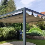 Wolnostojąca pergola rzymska Dragon o antracytowej, aluminiowej konstrukcji, umiejscowiona w bogato urządzonym ogrodzie. Zadaszenie wykonane z jasnobeżowej tkaniny jest rozsunięte, tworząc dekoracyjne fale między poprzecznymi profilami prowadzącymi. Solidna rama konstrukcji opiera się na pionowych, kwadratowych słupach, pod którymi znajduje się okrągły plac wyłożony jasną kostką brukową. W tle, bezpośrednio za pergolą, widoczny jest czarny grill gazowy ustawiony na tle gęstych, zielonych krzewów i roślin ozdobnych. Po prawej stronie kompozycji znajduje się półkolisty mur oporowy z kamienia, za którym rośnie niski, rozłożysty iglak. Dalszy plan wypełnia zadbany trawnik, wysokie drzewa iglaste oraz fragmenty sąsiednich budynków z dachami pokrytymi czerwoną dachówką. Zdjęcie wykonane w świetle dziennym, przy lekko zachmurzonym, jasnym niebie, co podkreśla elegancję i funkcjonalność ogrodowej strefy wypoczynku.