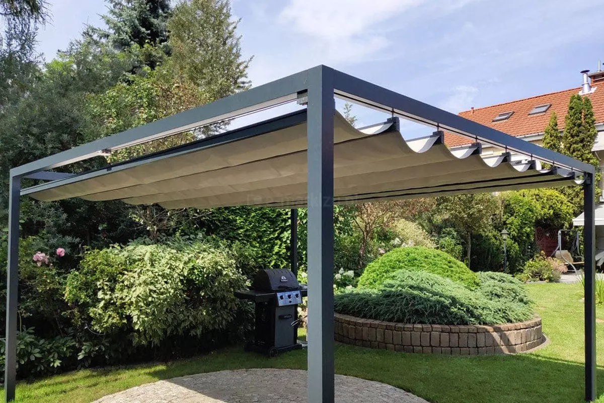 Wolnostojąca pergola rzymska Dragon o antracytowej, aluminiowej konstrukcji, umiejscowiona w bogato urządzonym ogrodzie. Zadaszenie wykonane z jasnobeżowej tkaniny jest rozsunięte, tworząc dekoracyjne fale między poprzecznymi profilami prowadzącymi. Solidna rama konstrukcji opiera się na pionowych, kwadratowych słupach, pod którymi znajduje się okrągły plac wyłożony jasną kostką brukową. W tle, bezpośrednio za pergolą, widoczny jest czarny grill gazowy ustawiony na tle gęstych, zielonych krzewów i roślin ozdobnych. Po prawej stronie kompozycji znajduje się półkolisty mur oporowy z kamienia, za którym rośnie niski, rozłożysty iglak. Dalszy plan wypełnia zadbany trawnik, wysokie drzewa iglaste oraz fragmenty sąsiednich budynków z dachami pokrytymi czerwoną dachówką. Zdjęcie wykonane w świetle dziennym, przy lekko zachmurzonym, jasnym niebie, co podkreśla elegancję i funkcjonalność ogrodowej strefy wypoczynku.