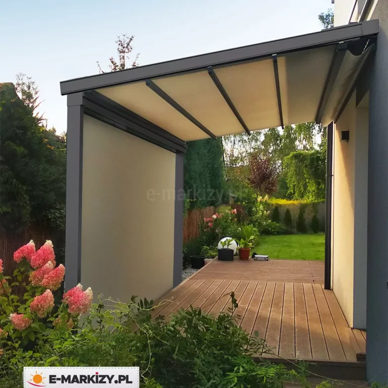 Nowoczesna pergola tarasowa o antracytowej, aluminiowej konstrukcji, zamontowana na wąskim tarasie nowoczesnego budynku mieszkalnego. Zadaszenie pergoli składa się z jasnego, kremowego poszycia materiałowego wspartego na równoległych profilach poprzecznych. Lewa strona konstrukcji została całkowicie osłonięta pionową przesłoną typu screen w kolorze dopasowanym do zadaszenia, co tworzy pełną ścianę boczną oddzielającą taras od sąsiedniej roślinności. Pergola jest zainstalowana nad tarasem wykonanym z brązowych desek kompozytowych, który płynnie przechodzi w zadbany zielony ogród. Po lewej stronie, na pierwszym planie, widoczne są kwitnące krzewy hortensji o różowo-białych kwiatach oraz gęsta, zielona roślinność. W głębi ogrodu znajdują się niskie nasadzenia, donice z roślinami oraz kula ogrodowa, a całość otacza gęsty żywopłot i drzewa. Zdjęcie wykonane w świetle dziennym, ukazujące przytulną i osłoniętą strefę relaksu zintegrowaną z bryłą domu.
